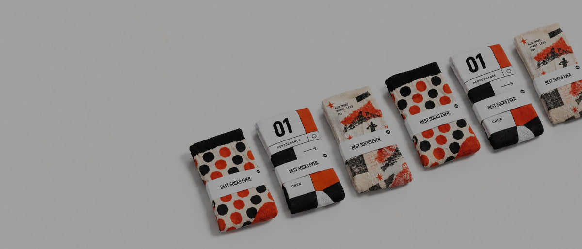 Six packaged pairs of patterned custom socks arranged diagonally on a clean white background. The socks feature various designs including orange and black polka dots, orange and black checkered patterns, and a collage print with orange accents.