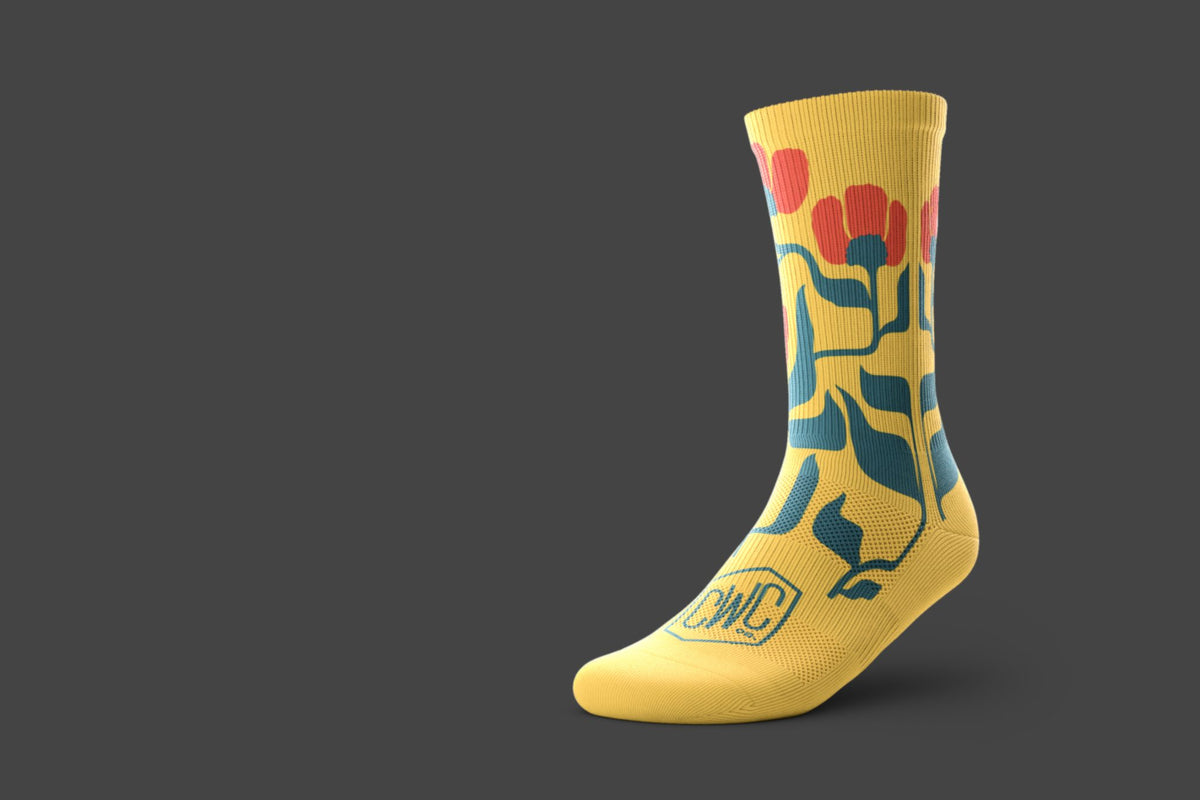 A single yellow performance crew sock with a stylized floral graphic in green and red, featuring a 'CWC' logo near the foot, shown on a dark gray background.