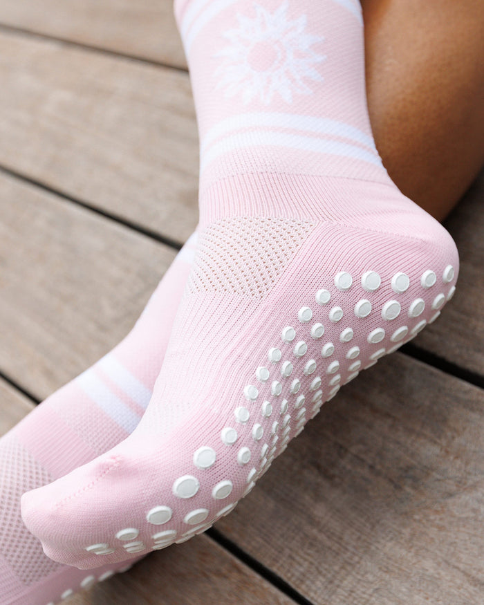 A close-up of a person's foot wearing a light pink grip sock with white grip dots on the sole and a white sun motif near the cuff.