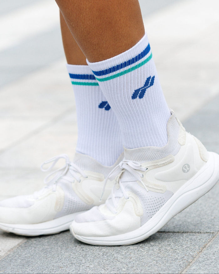 White athletic socks with blue and green stripes and a blue logo, worn with white sneakers on a paved outdoor surface.