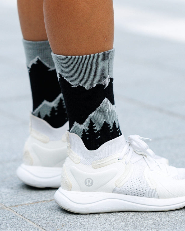 Gray crew socks with a black and gray mountain and pine tree pattern, worn with white athletic sneakers on a paved outdoor surface.