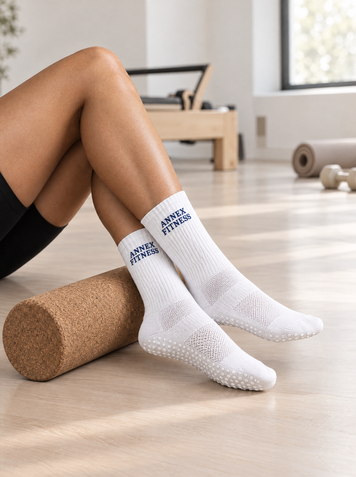 White grip crew socks with blue 'ANNEX FITNESS' text, worn by a person sitting on a cork foam roller in a bright fitness studio.