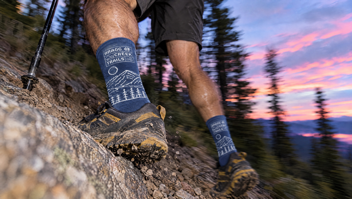 A close-up outdoor shot of a person hiking on a rocky trail, wearing blue socks with a white mountain and tree line graphic and the text 'BRAGG CREEK TRAILS.' paired with black and brown hiking boots.