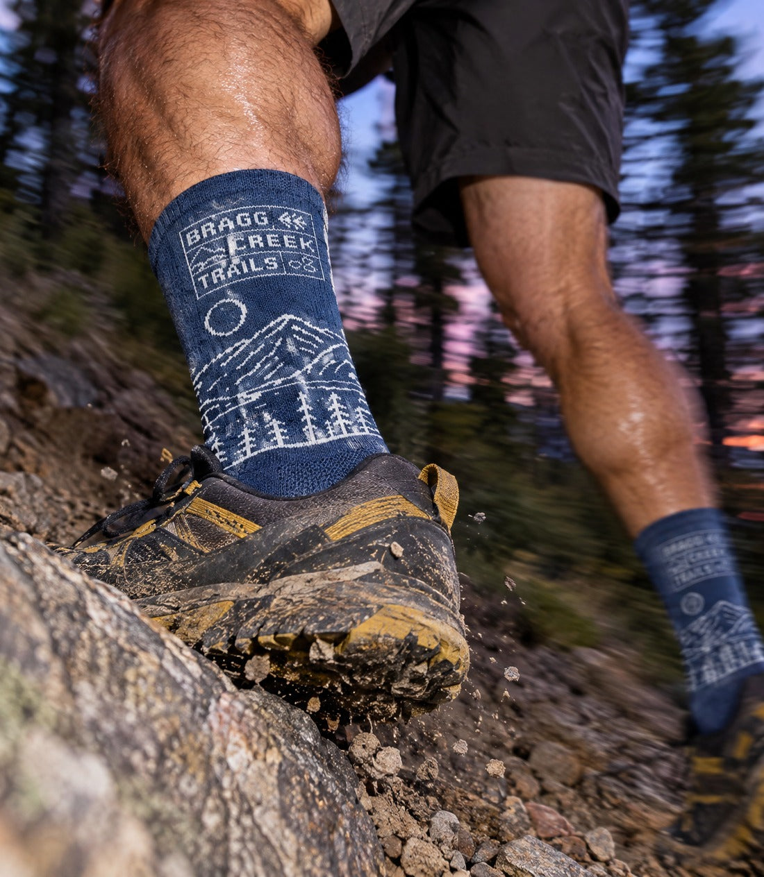 A close-up outdoor shot of a person hiking on a rocky trail, wearing blue socks with a white mountain and tree line graphic and the text 'BRAGG CREEK TRAILS.' paired with black and brown hiking boots.