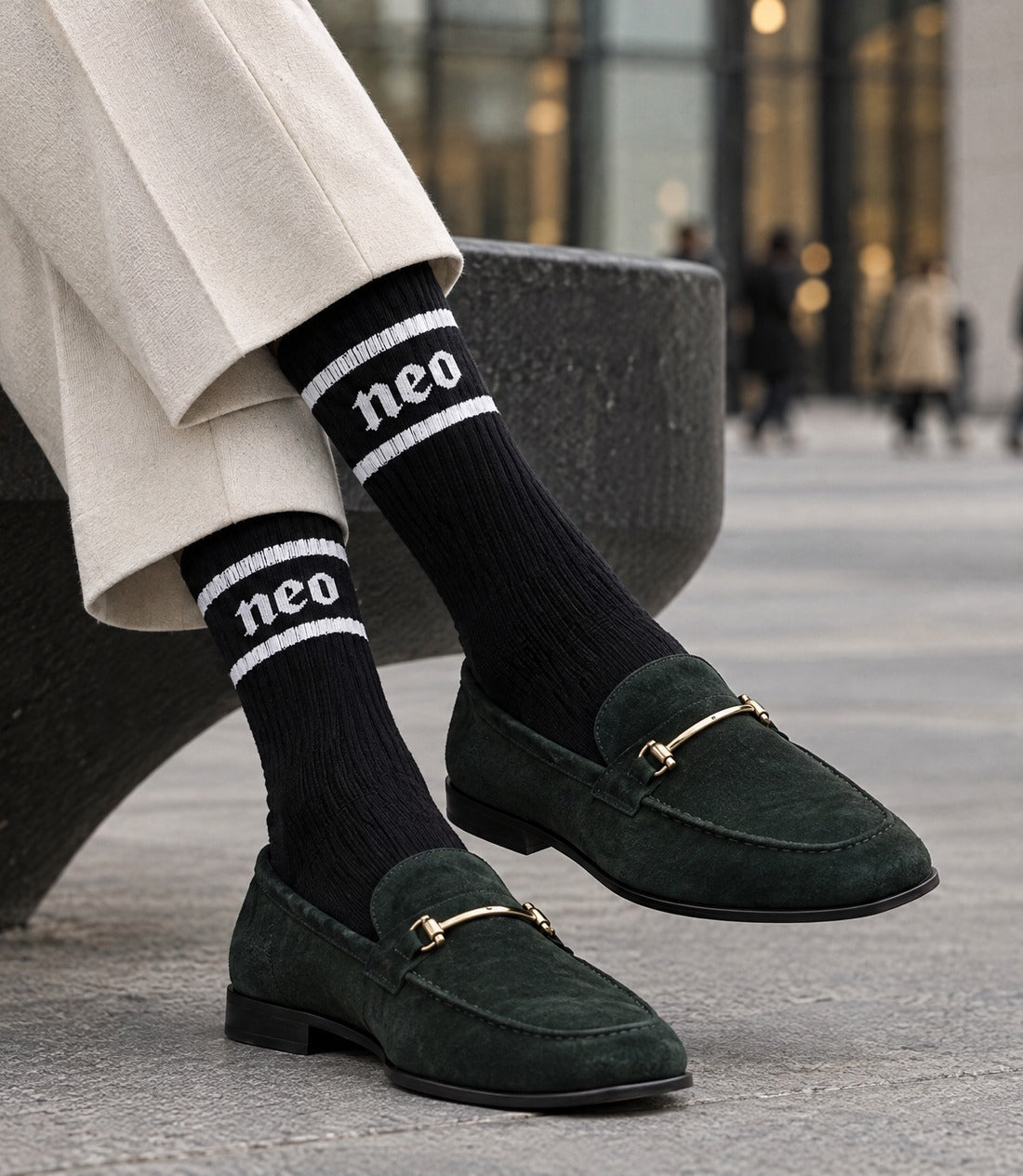 A person wearing dark green suede loafers with gold-tone horsebit details, paired with black cotton crew socks featuring white stripes and the word 'neo' near the top, seated on a dark stone bench in an outdoor urban setting.