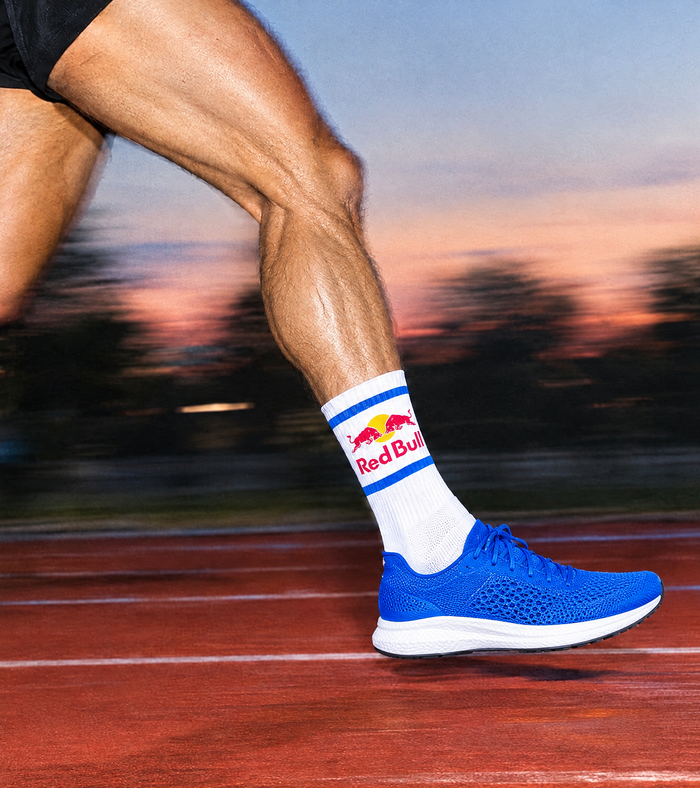 A close-up action shot of a runner's leg on a track during a race, wearing a white performance crew  athletic sock that reads 'Red Bull' and a bright blue running shoe against a blurred sunset background.