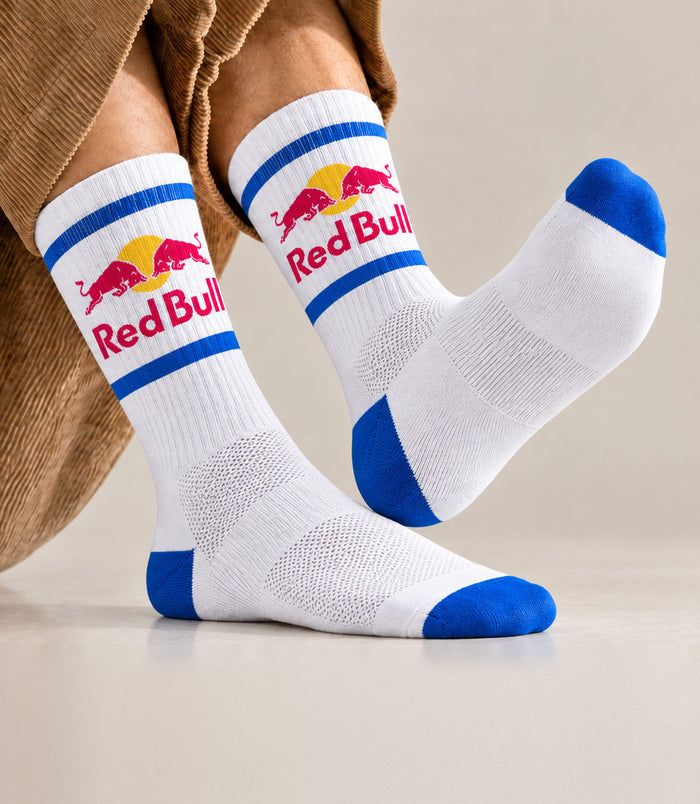 A person wearing Red Bull branded performance crew socks with blue toes, heels, and cuffs, paired with brown corduroy pants, shown against a neutral studio background.