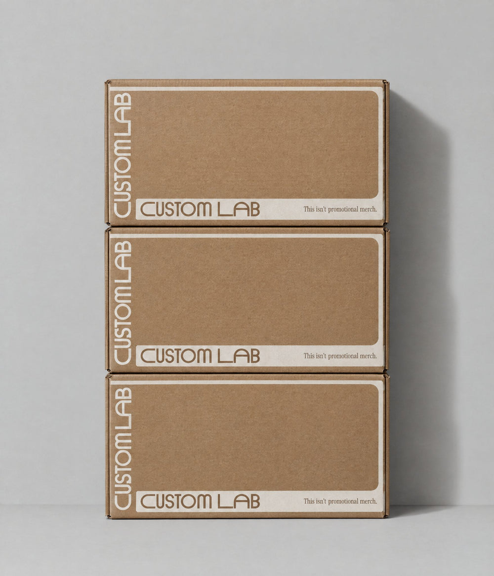Three stacked brown cardboard boxes with 'CUSTOM LAB' printed vertically on the sides, set against a neutral gray background.