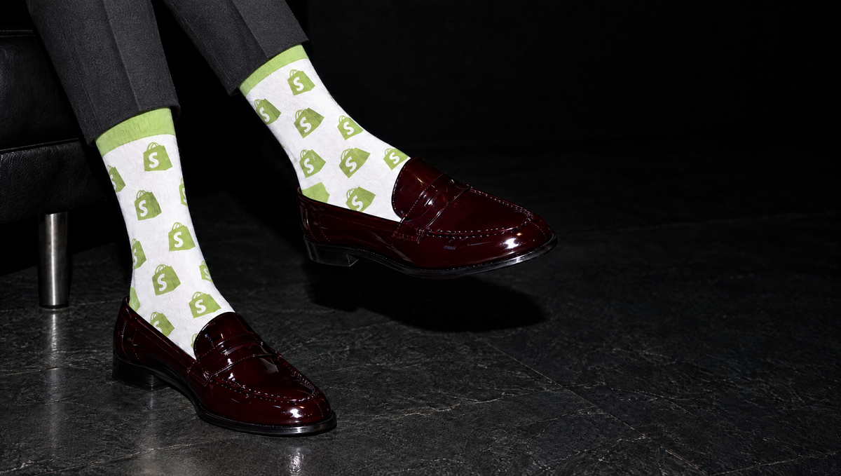 White dress socks featuring the 'Shopify' logo with a green shopping bag pattern featuring a white dollar sign , worn with dark burgundy patent leather penny loafers.