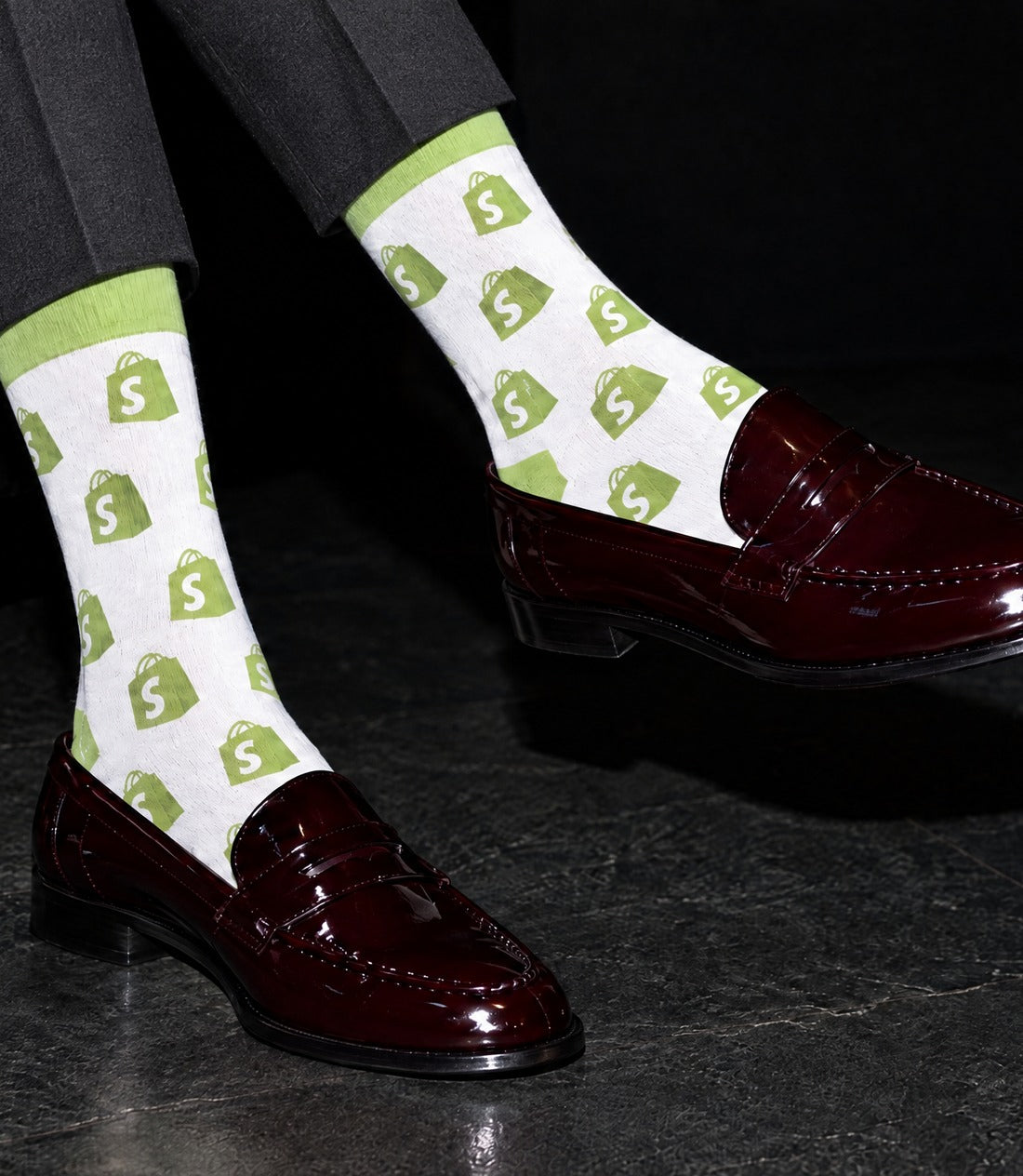 White dress socks featuring the 'Shopify' logo with a green shopping bag pattern featuring a white dollar sign , worn with dark burgundy patent leather penny loafers.