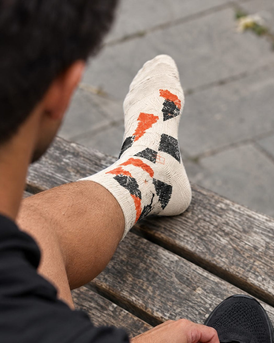 A person sitting on a wooden bench outdoors, tying the laces of a black athletic shoe while wearing patterned crew socks. The socks are off-white with orange and black geometric and graphic designs.