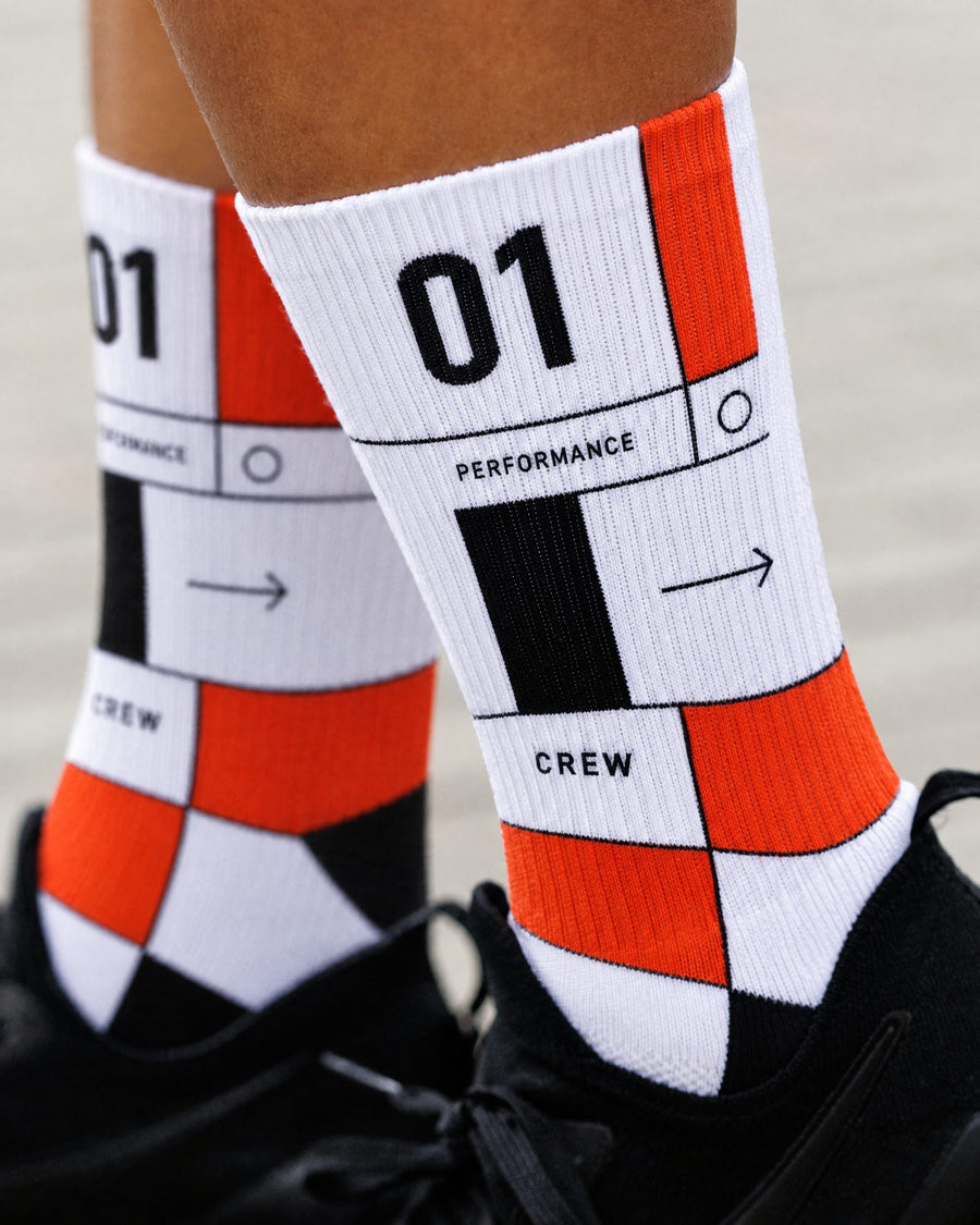 White performance crew socks with a bold orange and black pattern and printed text including '01', 'PERFORMANCE', and 'CREW', worn on feet with black sneakers.