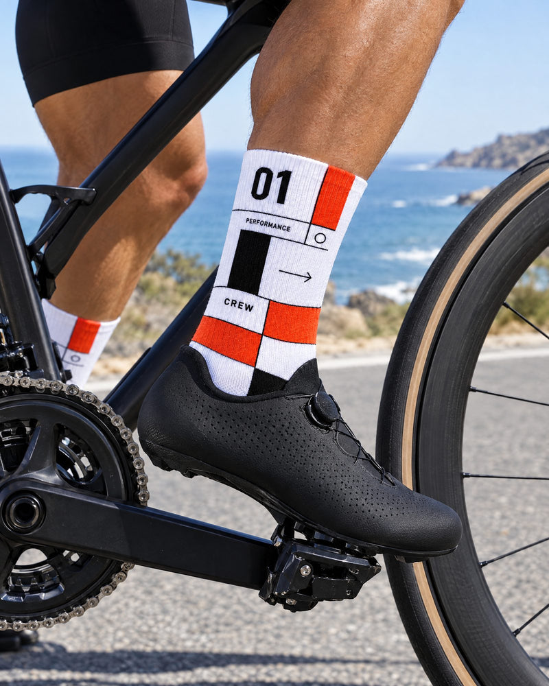 A close-up lifestyle shot of a cyclist's leg wearing a black cycling shoe and white crew socks with orange, black, and white graphic details, riding a black road bike on a coastal road with the ocean in the background.