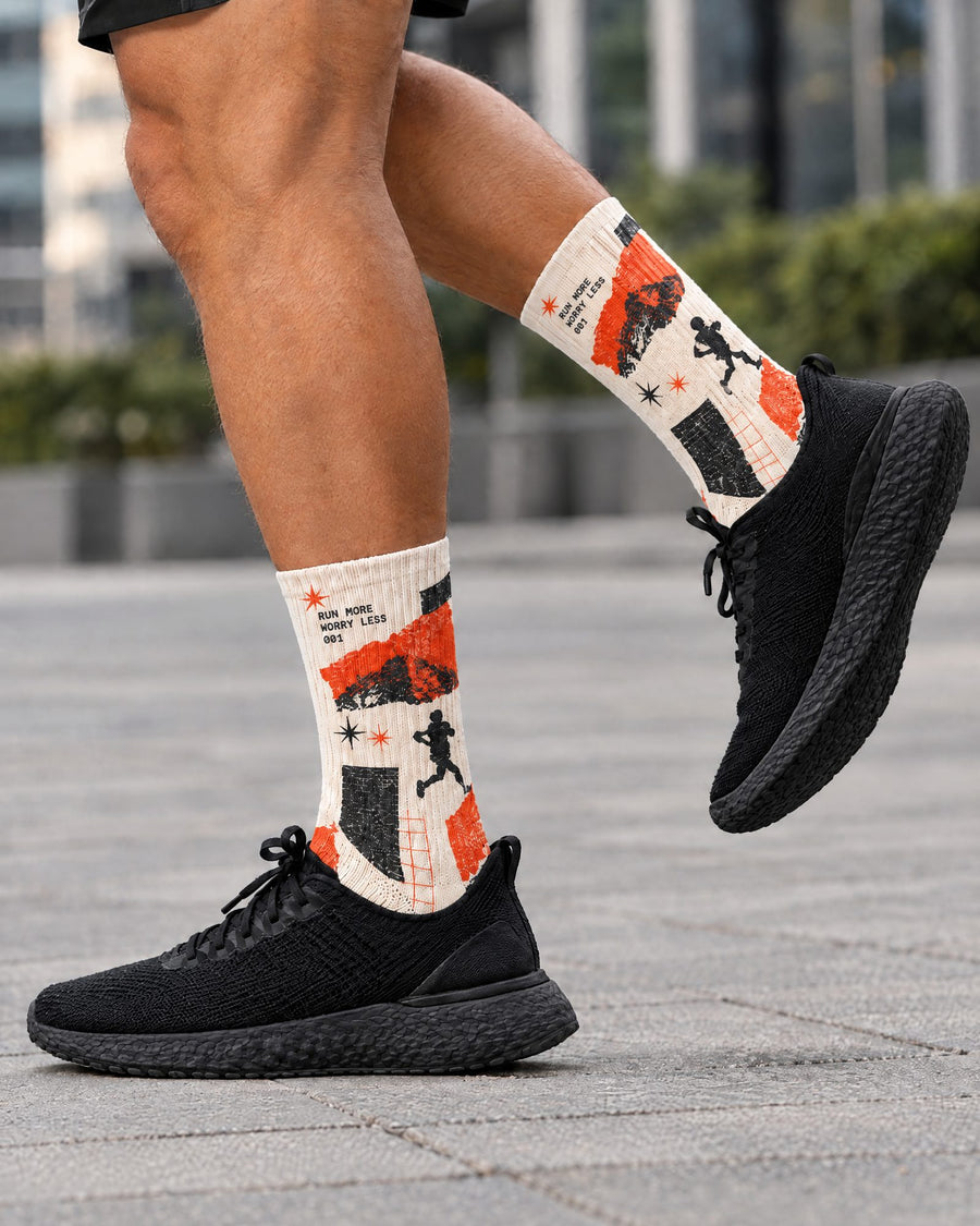 A person outdoors wearing black sneakers and custom performance crew socks with a bold orange, black, and off-white graphic print featuring running silhouettes and text.
