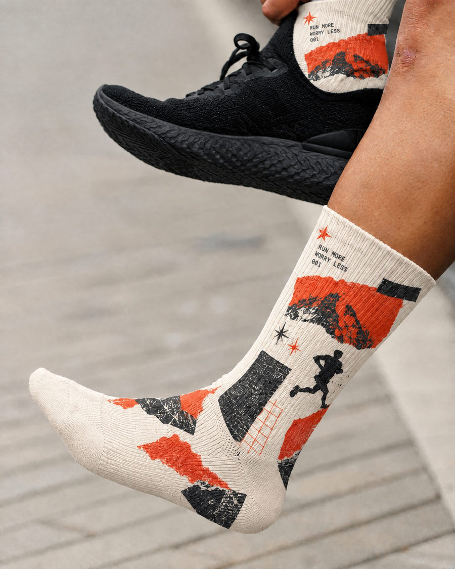 A person wearing patterned performance crew socks with a black running shoe, photographed outdoors on a paved surface. The socks are off-white with bold orange, black, and gray graphic shapes and small text, including 'RUN MORE WORRY LESS.'