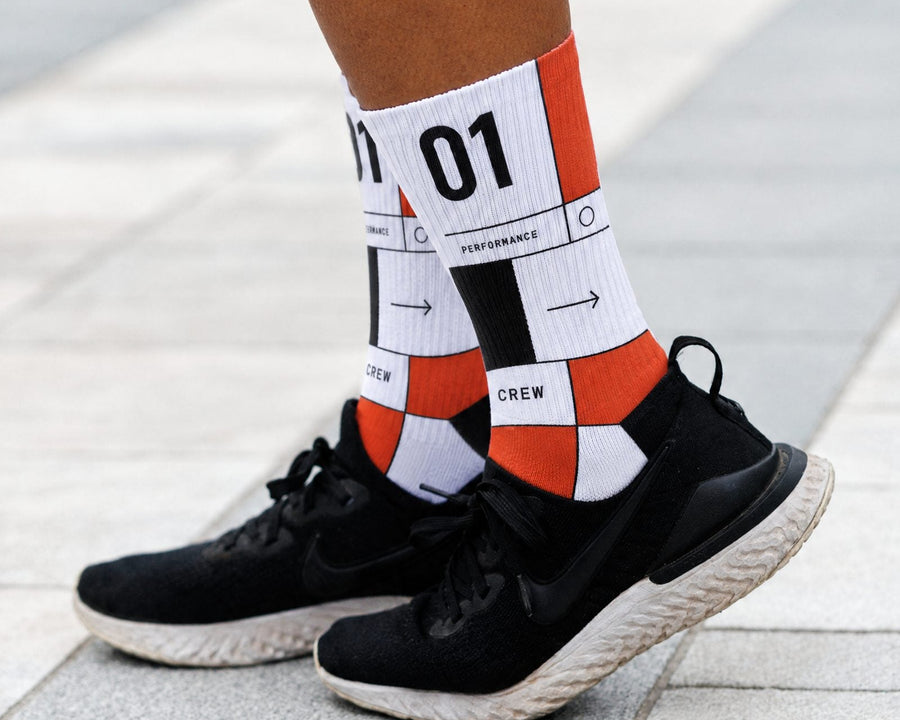 A close-up of a person's legs wearing white performance crew socks with an orange geometric pattern and black text, paired with black running shoes on a gray paved surface.