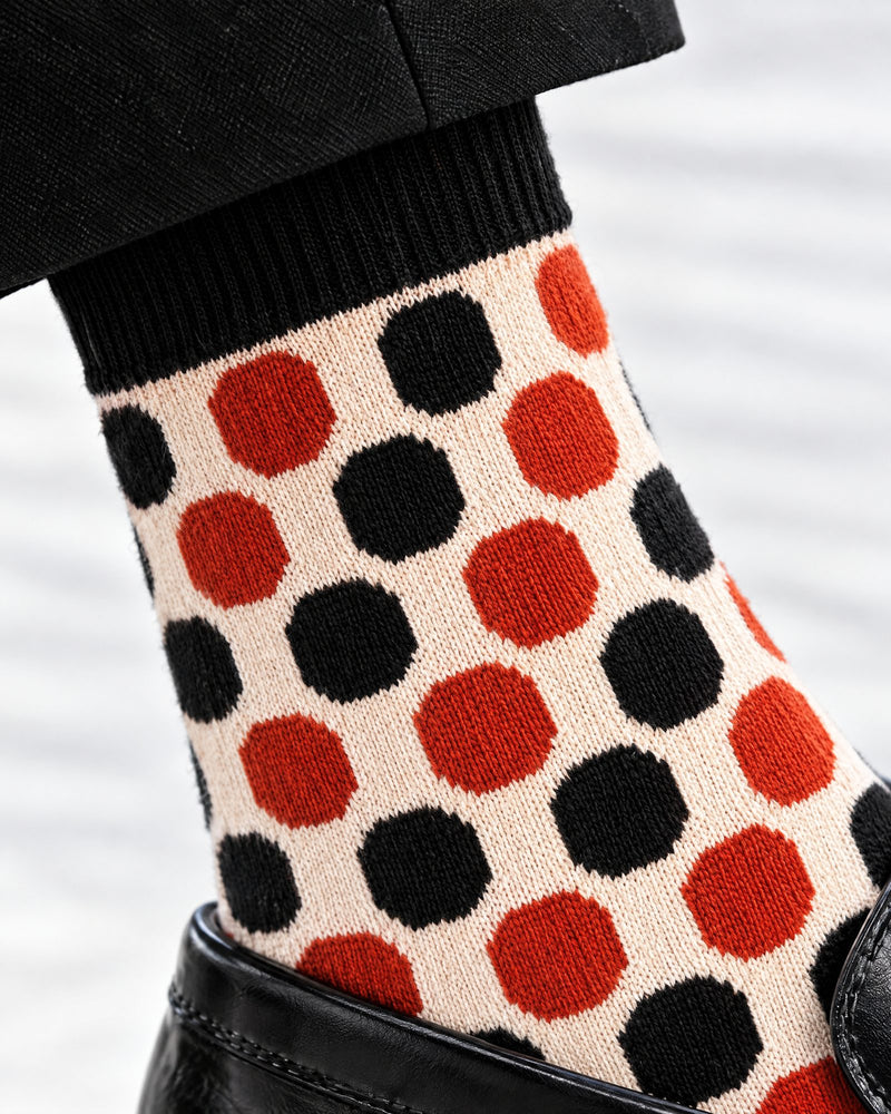 A close-up of a patterned knit sock worn on a foot, featuring large alternating orange and black polka dots on a warm cream background, paired with a black cuff and black shoe.