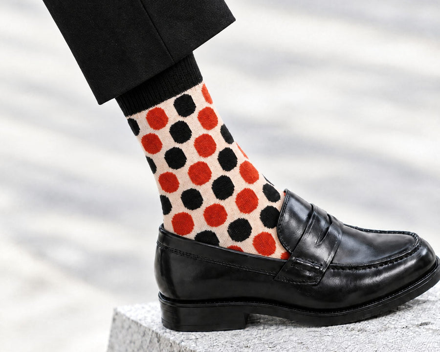 A close-up of a person's lower leg wearing black trousers, a cream dress sock with a bold oranage and black polka dot pattern, and a black leather penny loafer, standing on a stone block.