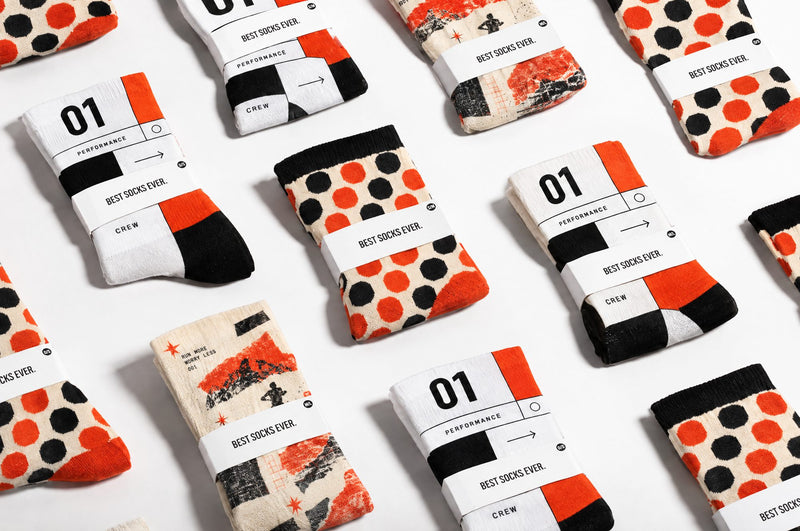 Multiple pairs of custom socks arranged in a flat-lay pattern on a white background. Each pair is wrapped in a white paper band labeled 'BEST SOCKS EVER'. Showing different color and pattern designs including orange and black polka dots, black and white stripes, and abstract orange, black, and white prints.
