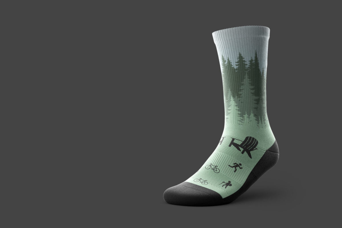 A single crew sock shown on a dark gray background, featuring a light mint green and dark green forest pattern with black accents on the toe, heel, and sole. It also features a chair, bikes, and people hiking. 