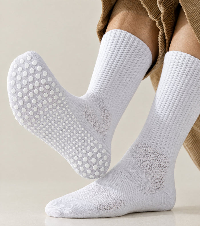 White ribbed grip socks worn on a person's feet, with one sock turned to show the textured non-slip sole. The person is wearing brown corduroy pants against a light beige background.