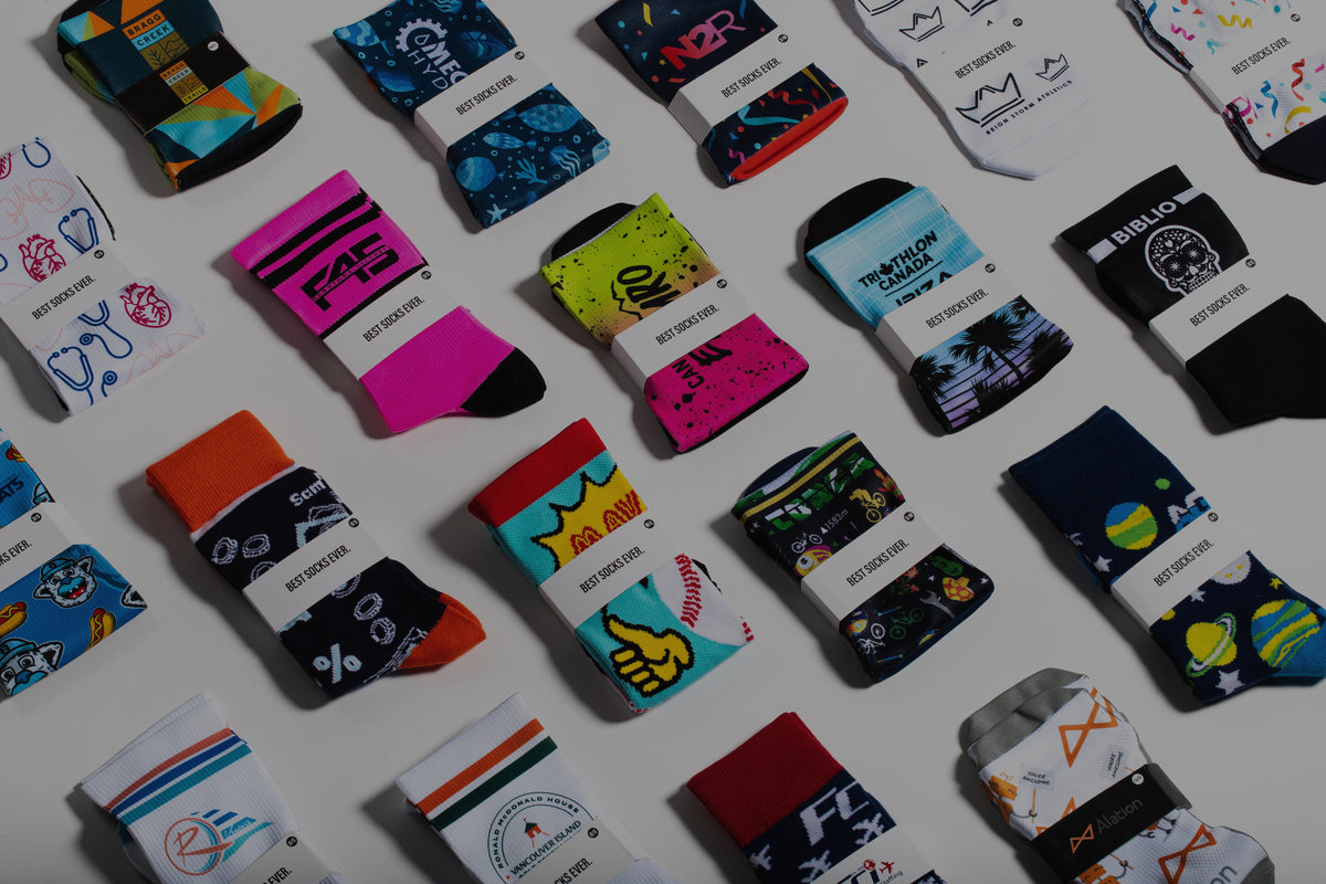 A flat-lay display of multiple pairs of colorful patterned custom socks from different brands arranged diagonally on a white background. Each pair is wrapped in a white paper band with the words 'BEST SOCKS EVER' visible.