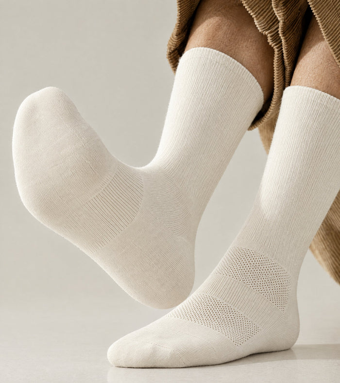 A close-up of a person's feet wearing off-white merino wool crew socks, with the legs and hem of a brown corduroy trousers visible against a clean, light beige studio background.