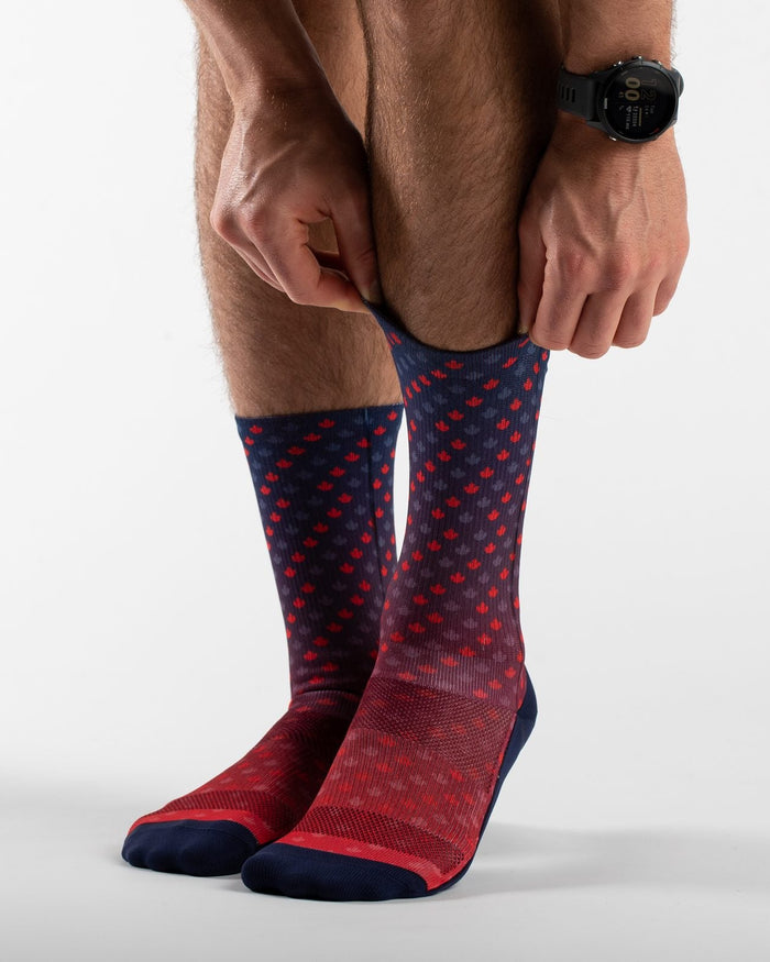 A close-up of a person's lower legs and feet as they pull up a pair of red and navy blue patterned crew socks against a clean white studio background.