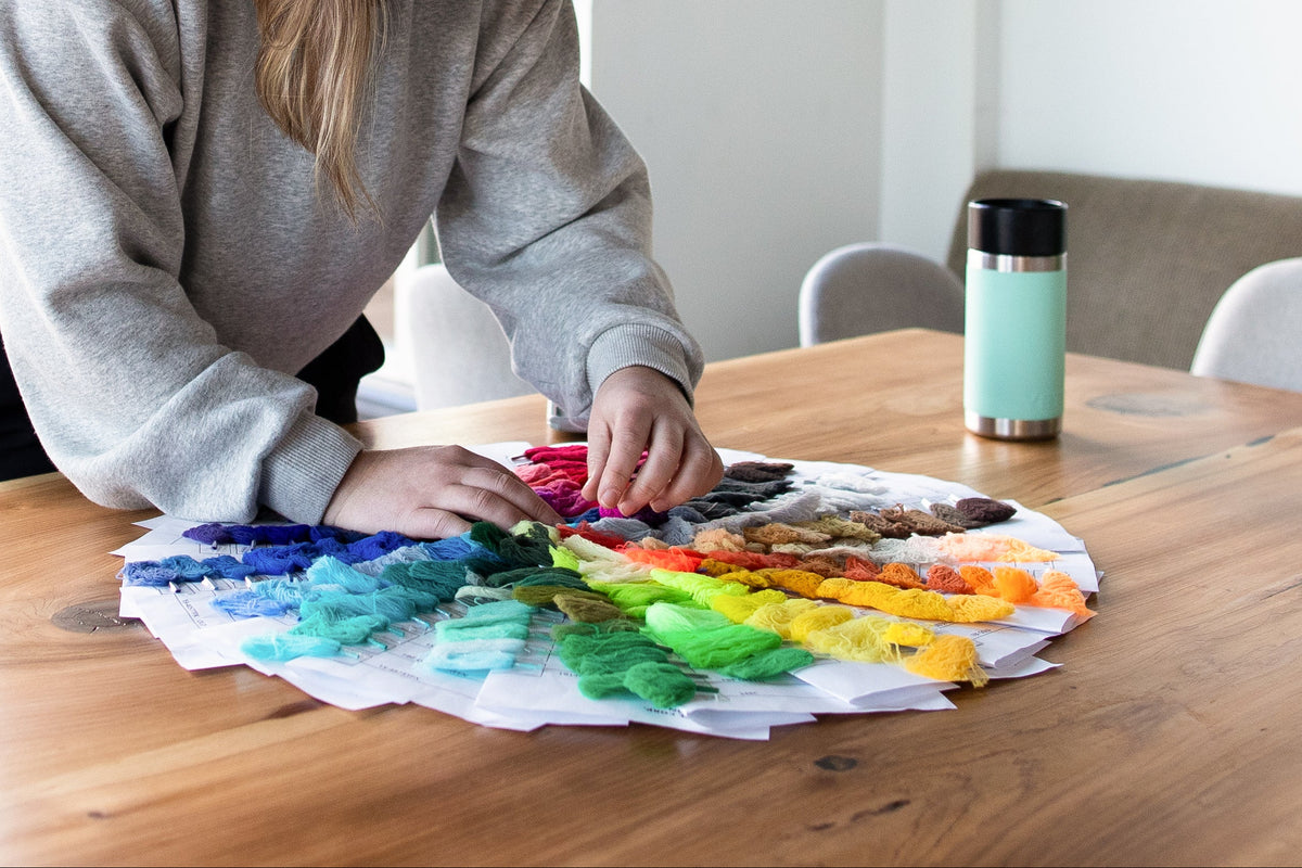 A woman in a gray sweatshirt leans over a wooden table, arranging colorful circular swatches of yarn a large rainbow wheel, modern indoor setting.